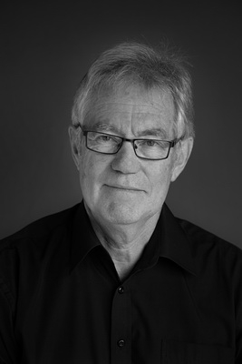 Elderly man in black shirt with serious expression, neutral background.